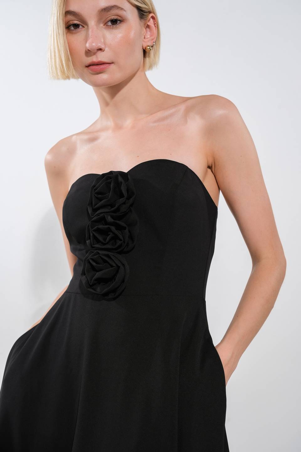 A woman is wearing a strapless black dress featuring three fabric roses on the front. She stands against a light-colored background, presenting a poised expression and subtly holding her hands in pockets.