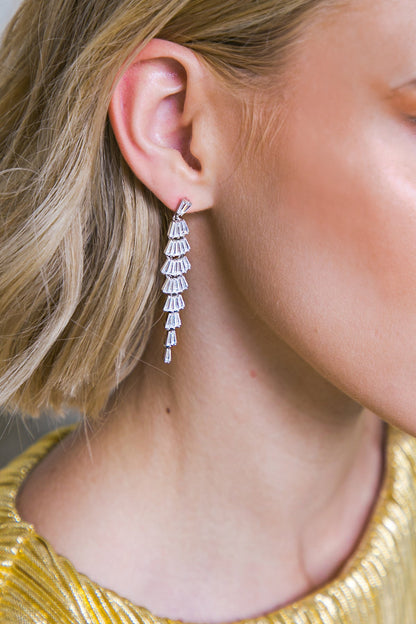 A diamond earring dangles from a woman's ear. She has short, blonde hair, and is wearing a shiny gold top, suggesting a formal or elegant setting.