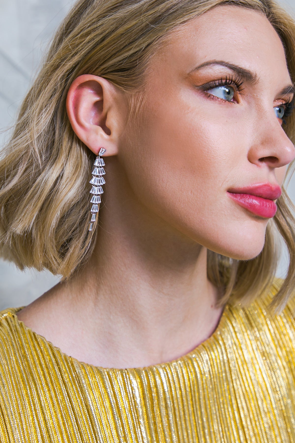 A woman with short, blonde hair wears a sparkling, cascading silver earring. She has a makeup look featuring bold lips and highlights, set against a neutral background.