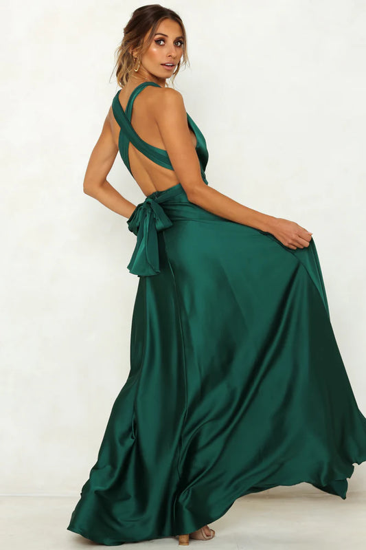 A woman dressed in a flowing, deep green gown turns to face the camera, showcasing the back design and bow detail, set against a neutral background.