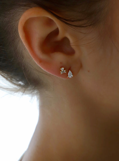 A close-up of an ear adorned with two different earrings, one a small diamond stud and the other a pear-shaped diamond, against a softly blurred light background.