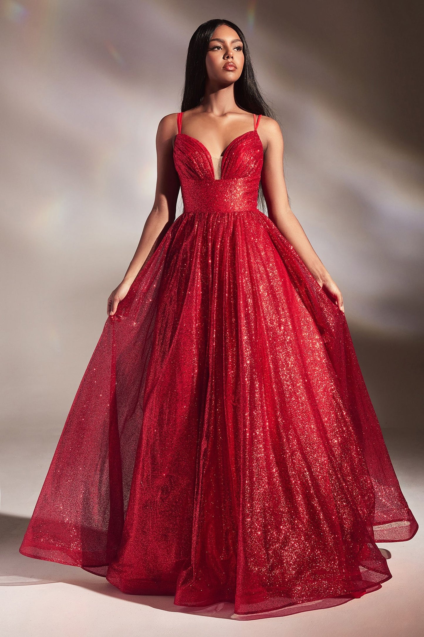 A woman stands confidently in a shimmering red gown with a fitted bodice and flowing skirt. The background is softly lit, enhancing the gown's sparkle and her poised expression.