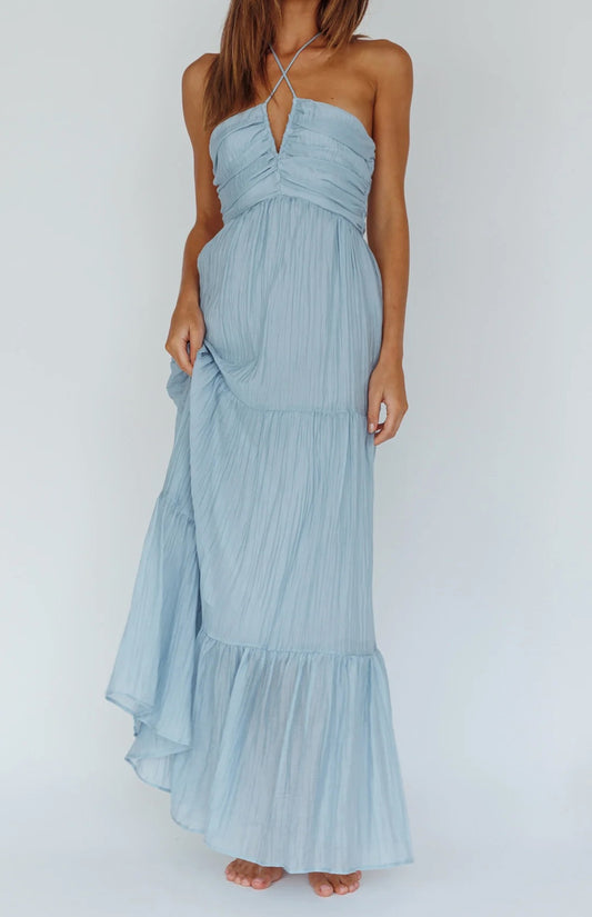 A woman stands wearing a flowy, light blue maxi dress with a halter neck and a gathered bodice, showcasing a relaxed pose against a plain background.