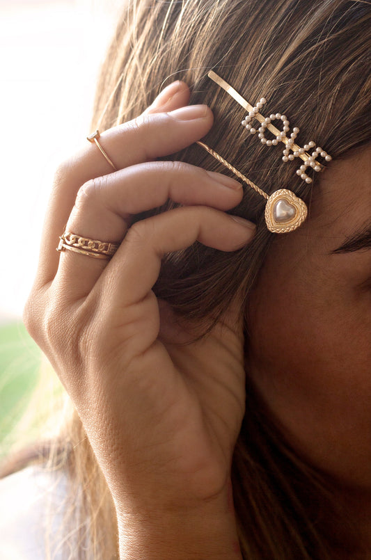 A hand adorned with delicate gold rings holds hair, featuring decorative hairpins with "LOVE" in pearls and a heart-shaped pin. The background is softly blurred, hinting at a warm, inviting space.