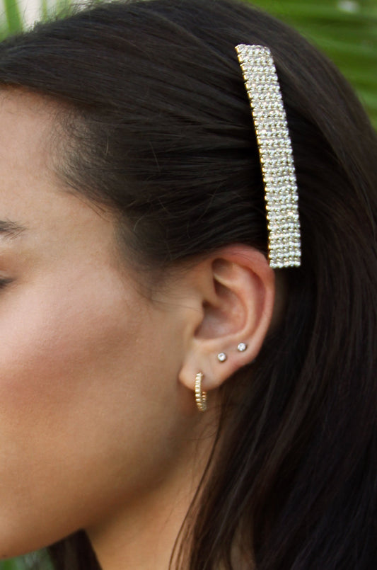 A hair accessory adorned with rhinestones is secured in dark, wavy hair next to a womanâ€™s ear, showcasing multiple earrings, while blurred greenery serves as the background.