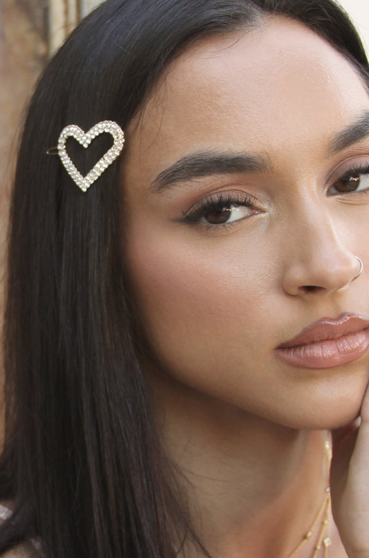 A woman with long, dark hair poses, showcasing a sparkling heart-shaped hair clip. She has natural makeup, softly lit skin, and an elegant expression against a blurred background.