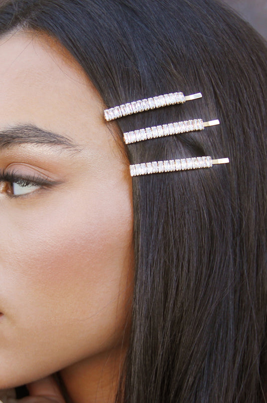 A close-up of a woman's profile features three shiny hairpins securing straight, dark hair against her head. The background is blurred, highlighting her makeup and the hair accessories.