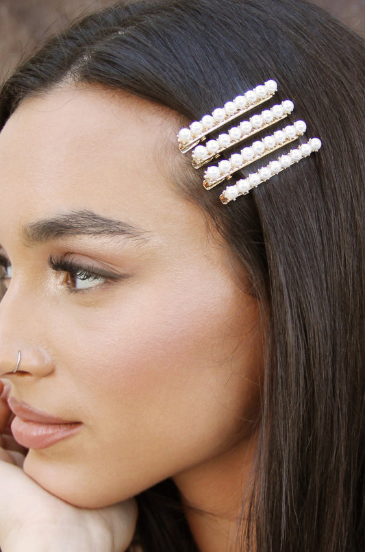 A hair clip with four rows of pearls is placed in long, dark hair. A woman rests her chin on her hand, showing a soft, natural makeup look against a neutral background.