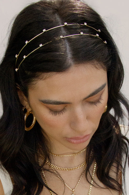 A woman with dark, wavy hair wears a decorative headband with small beads while looking downward. She has multiple gold necklaces and large hoop earrings, with a neutral background.