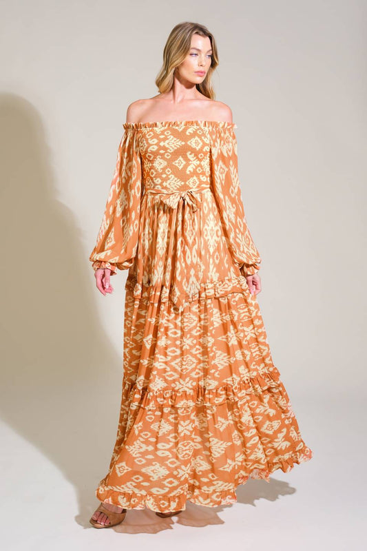 A model wears a flowing, off-the-shoulder, patterned maxi dress with long sleeves, standing against a light beige background, exuding a relaxed, elegant vibe.