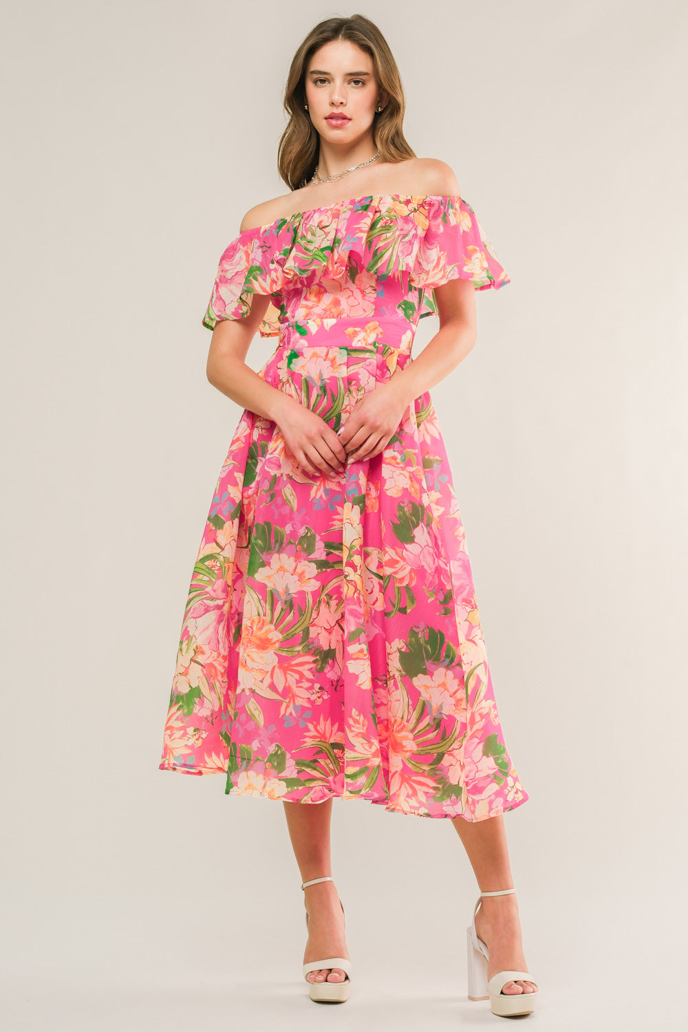 A woman stands confidently wearing a vibrant pink off-the-shoulder dress adorned with floral patterns. She poses with hands clasped, set against a soft, neutral background.