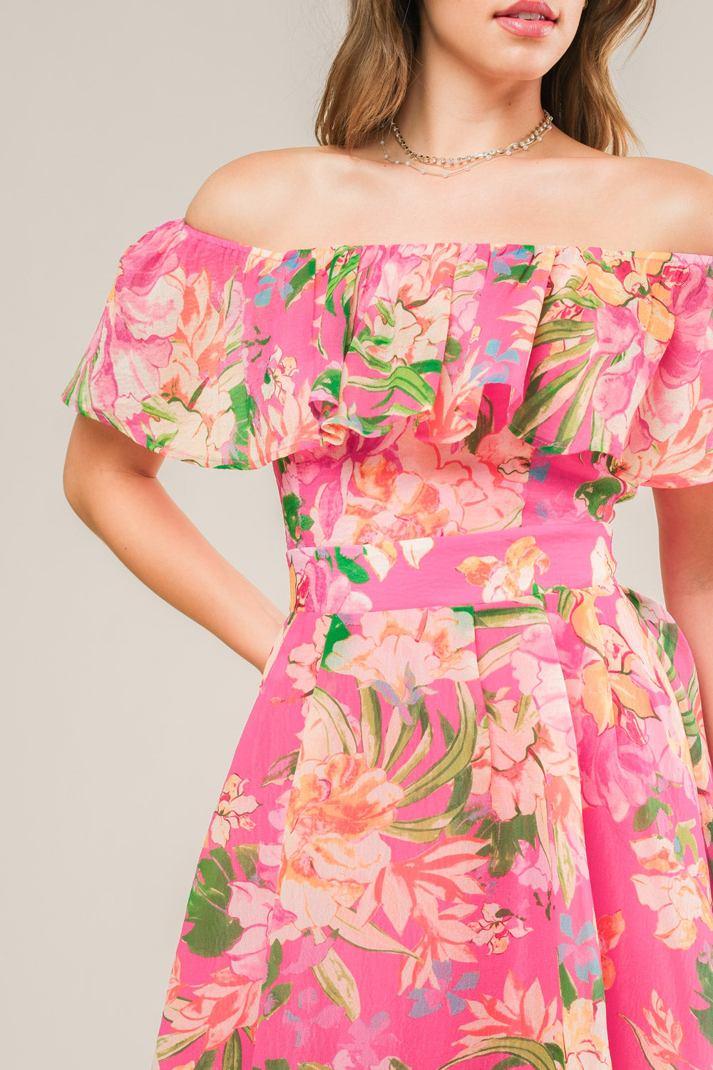 A floral dress in vibrant pink features an off-the-shoulder neckline and ruffled sleeves. The dress has a fitted waist and is set against a neutral background, highlighting its colorful design.