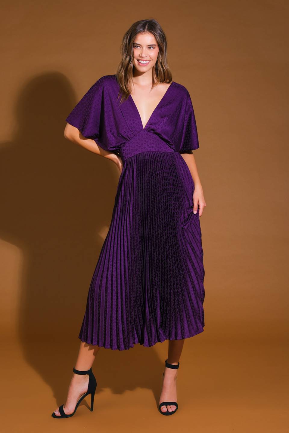 A model stands confidently in a flowing purple dress with a deep neckline and pleated skirt, showcasing elegance against a warm brown background, paired with black high-heeled sandals.