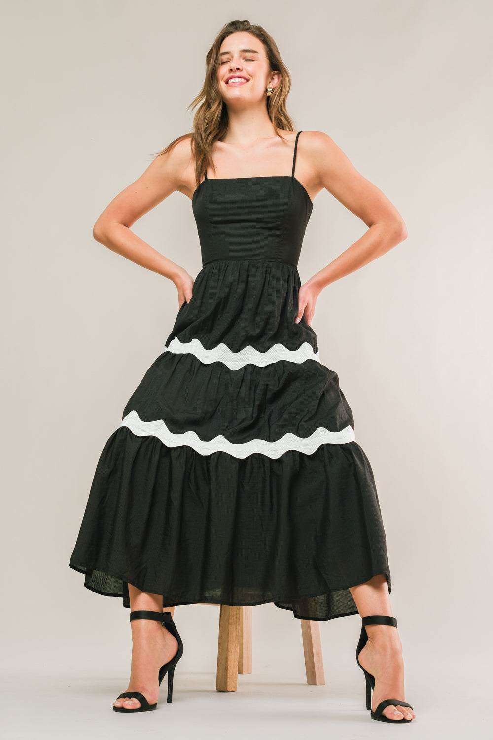 A woman smiles confidently while standing on a wooden stool, wearing a black, tiered dress with white ruffled accents and black high-heeled sandals, set against a neutral background.