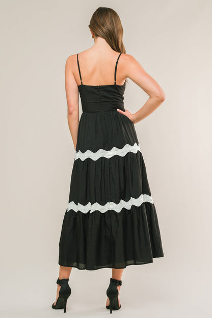 A woman is seen from the back, wearing a black, tiered maxi dress with white ruffles along the seams, paired with black high-heeled shoes, standing against a neutral background.