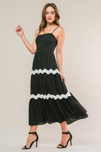 A woman in a black maxi dress with white ruffles stands gracefully, one hand raised, in a neutral-toned setting. Her long hair falls loosely, and she wears black high-heeled sandals.