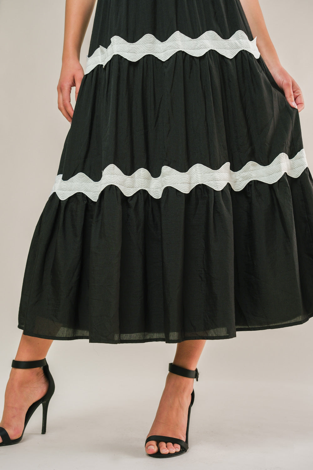 A black, tiered dress with white wavy trim is being worn by a person. The individual stands in black high-heeled sandals, showcasing the dress against a neutral background.