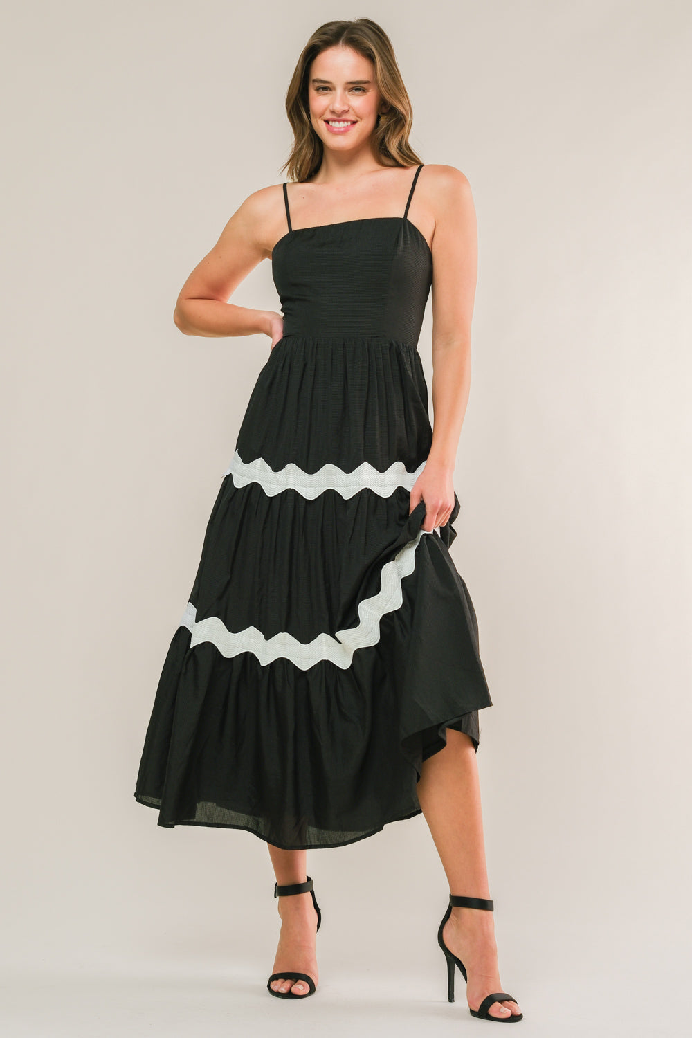 A woman stands confidently in a black, tiered maxi dress with white wavy accents. She poses with one hand on her hip and a smile, in a neutral studio background.