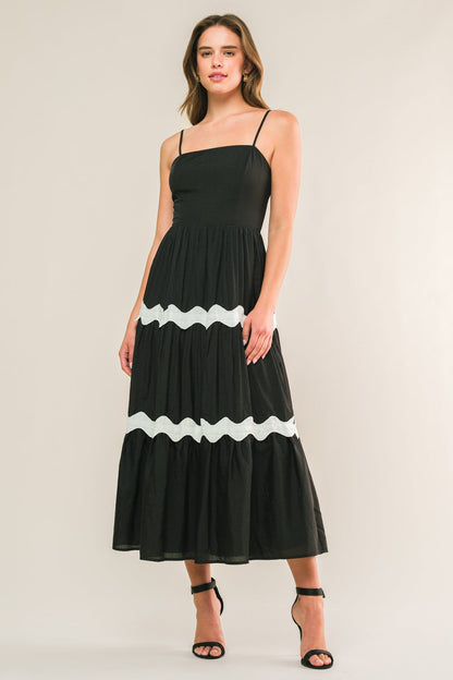 A woman stands wearing a black, sleeveless, tiered maxi dress with white wavy detailing. She smiles softly, against a plain, light-colored background, showcasing the dress's elegant design.