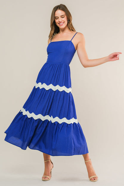 A woman in a blue, tiered sundress with white scalloped accents smiles and gracefully twirls, standing against a neutral background that emphasizes her joyful demeanor and outfit.