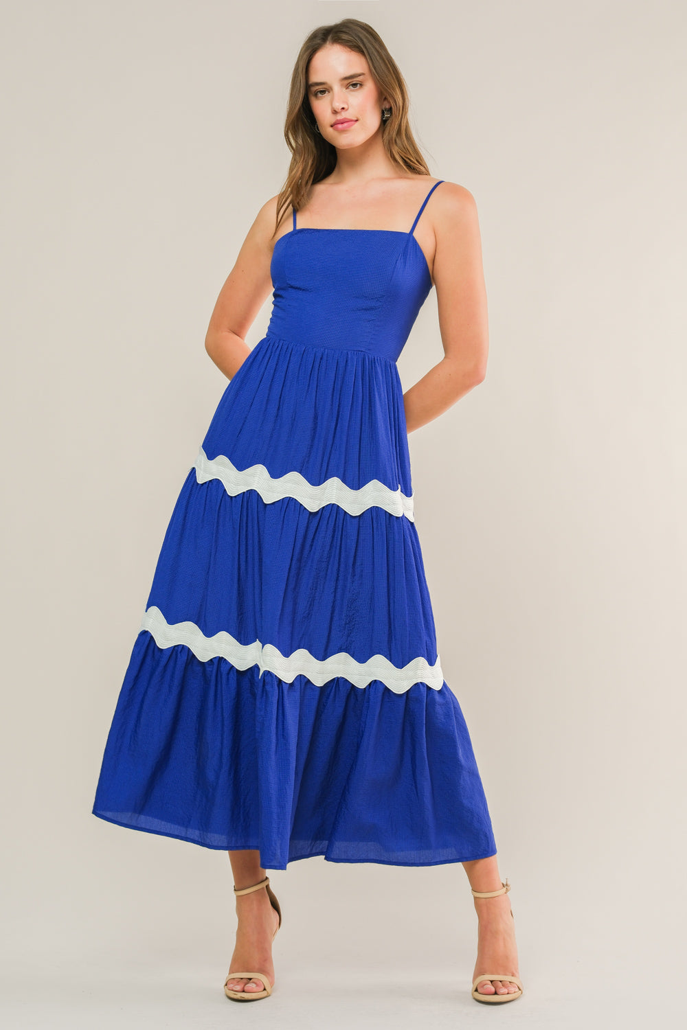 A young woman stands confidently wearing a vibrant blue maxi dress with white scalloped trim. She's in a neutral background, showcasing the dress's flowing silhouette and her casual yet elegant pose.