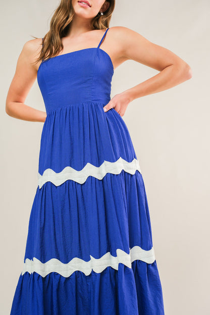 A woman poses in a vibrant blue maxi dress with white scalloped trim, standing against a neutral background. Her hand is on her hip, highlighting the dress's flowing design.