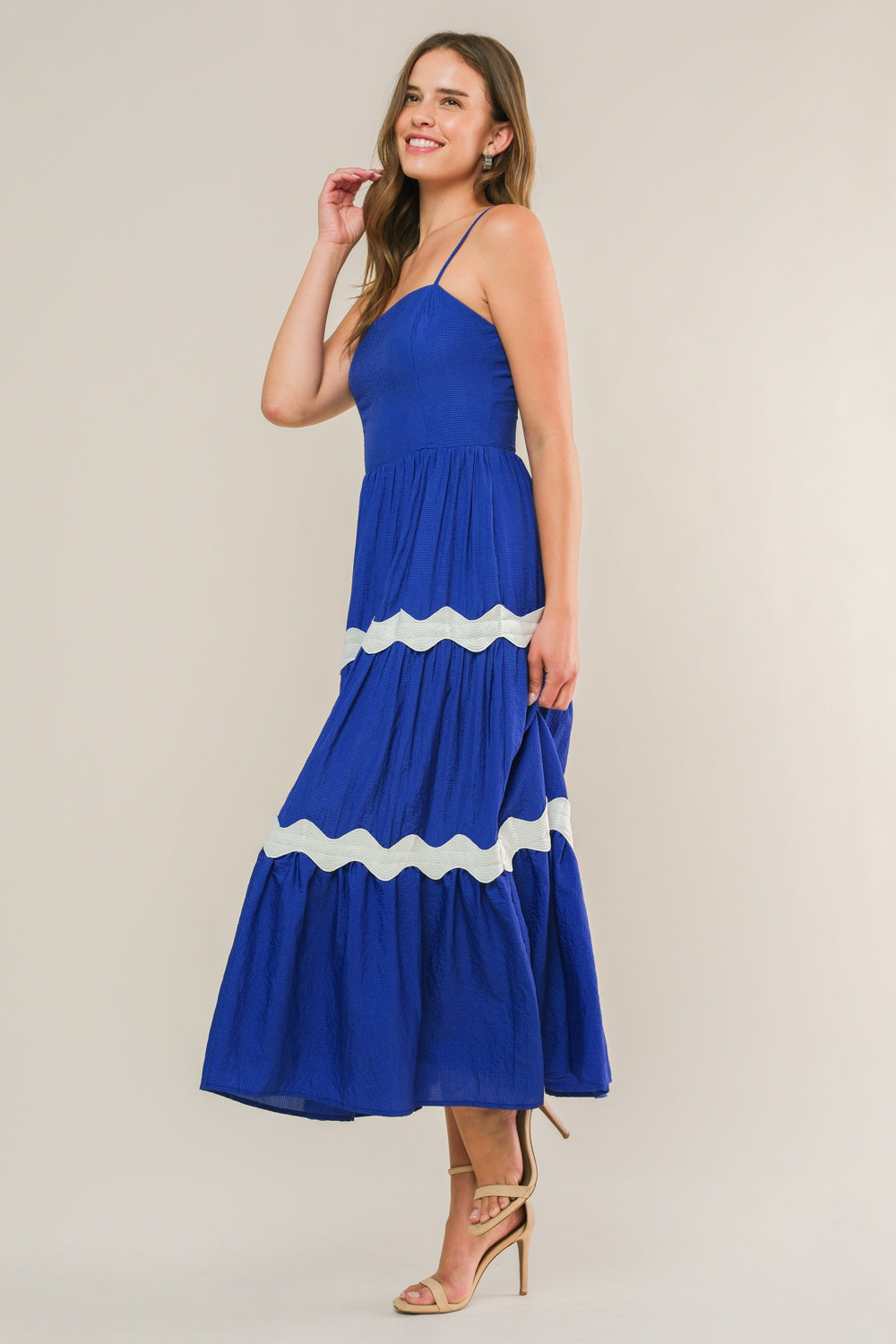 A woman stands gracefully, wearing a vibrant blue maxi dress with white wavy trim. She lightly touches her hair and smiles, set against a plain light backdrop.