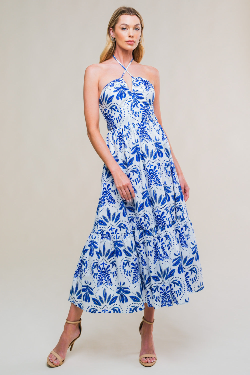 A woman poses confidently in a blue and white floral-patterned maxi dress, standing against a neutral background that enhances the dress's colors and designs. Sandals complement her look.