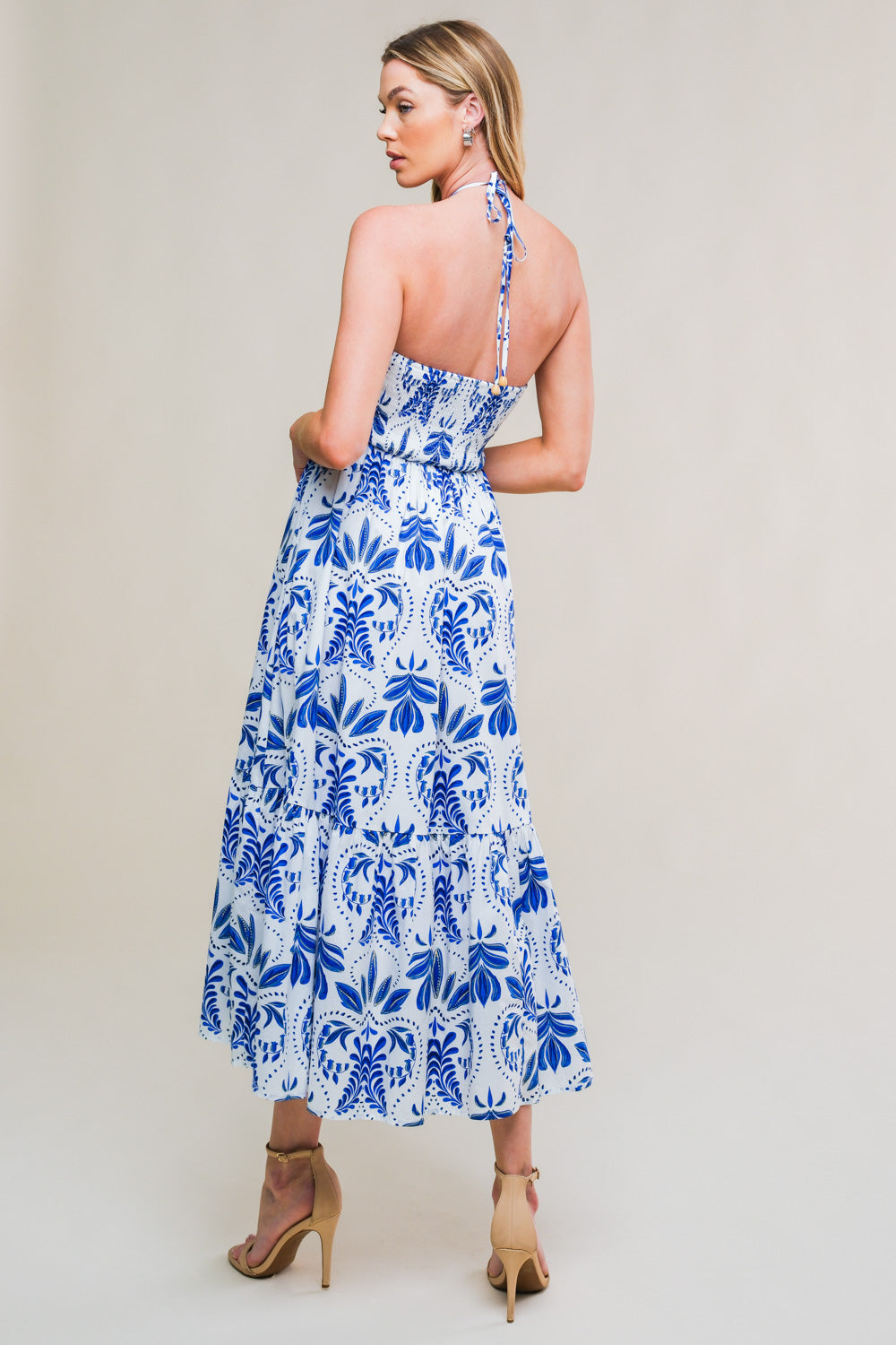 A woman stands in profile wearing a blue and white floral maxi dress that ties at the back. She has long hair and is paired with beige high-heeled sandals against a light background.