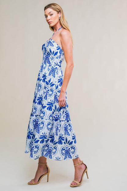 A woman stands elegantly in a long, patterned dress featuring blue floral designs against a white background. She wears nude high-heeled sandals and poses with a confident expression. The background is simple and light.