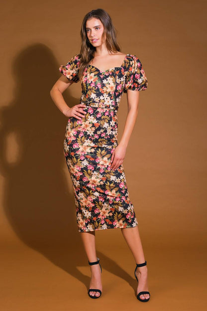 A woman poses confidently in a floral-patterned dress with puffed sleeves, showcasing her outfit against a warm brown background. She wears black strappy heels and smiles slightly.