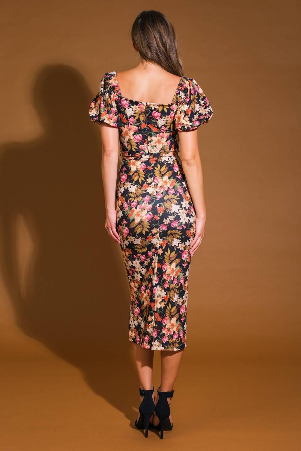 A woman stands with her back to the viewer, wearing a form-fitting, floral-patterned dress with puffed sleeves. The background is a warm brown, enhancing the vibrant colors of the dress.