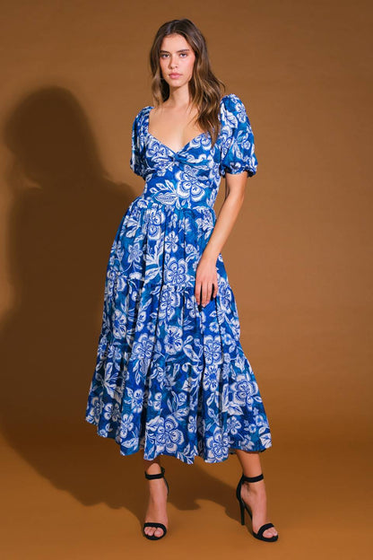 A woman stands confidently in a blue floral dress with puffed sleeves, showcasing its layered skirt. The background is a warm brown, enhancing the dress's vibrant colors.