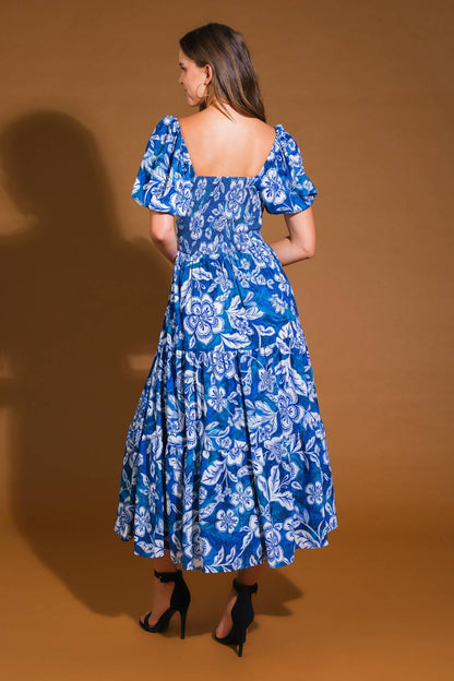 A woman stands with her back to the viewer, wearing a blue floral dress with puffed sleeves and a tiered skirt, against a warm brown backdrop. She wears black high-heeled shoes.