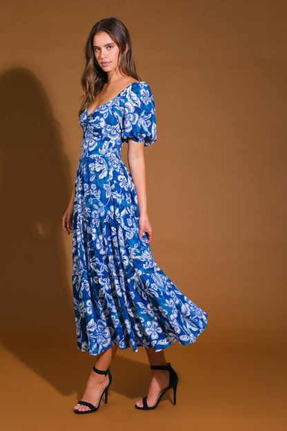 A woman wears a blue floral dress with puffed sleeves, posing confidently. Her heels accentuate her legs. The background is a warm brown, enhancing the dress's vibrant colors.