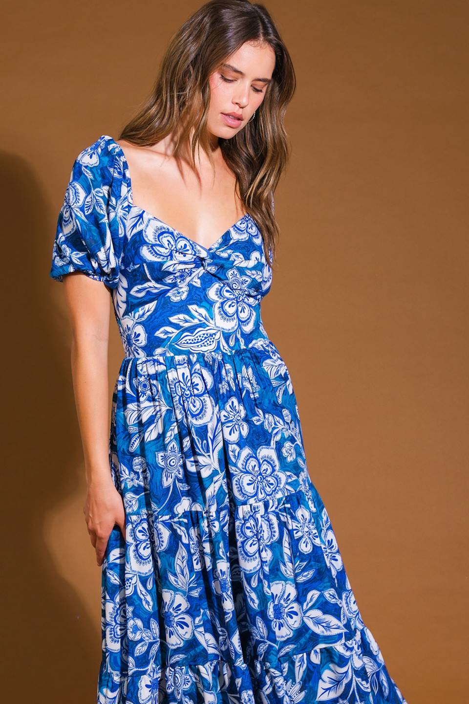 A woman wears a blue floral dress, gently looking down with a soft expression. The background is a warm brown, highlighting the dress's vibrant patterns.