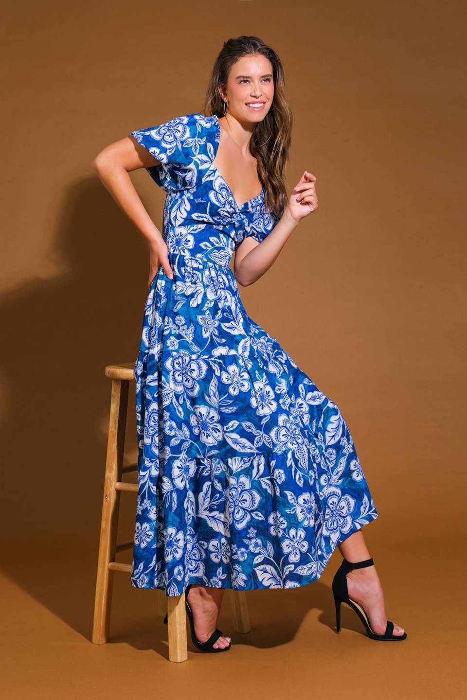 A woman poses on a wooden stool, wearing a blue floral maxi dress and black heels. She smiles, exuding a cheerful vibe against a warm brown backdrop.