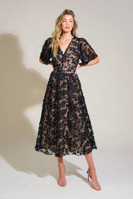 A woman stands confidently, wearing a black floral lace dress with puffed sleeves and a fitted bodice. She poses against a light neutral background, highlighting the intricate patterns of the garment.