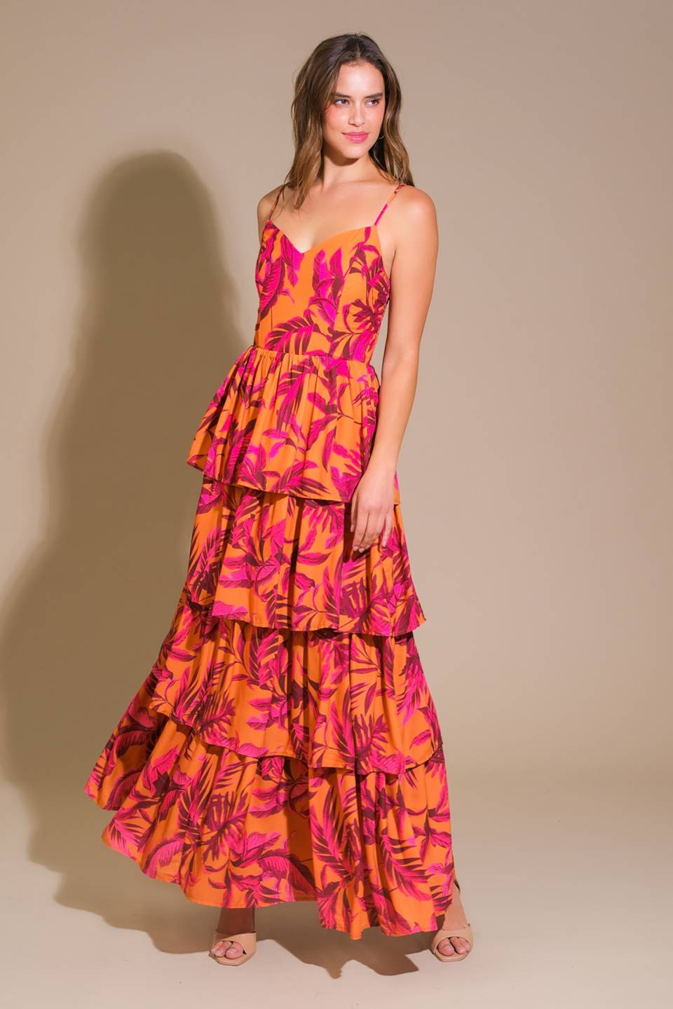 A model stands elegantly, wearing a vibrant, tiered maxi dress featuring bold orange fabric with pink leaf patterns. The backdrop is a neutral beige, enhancing the dress's colors.