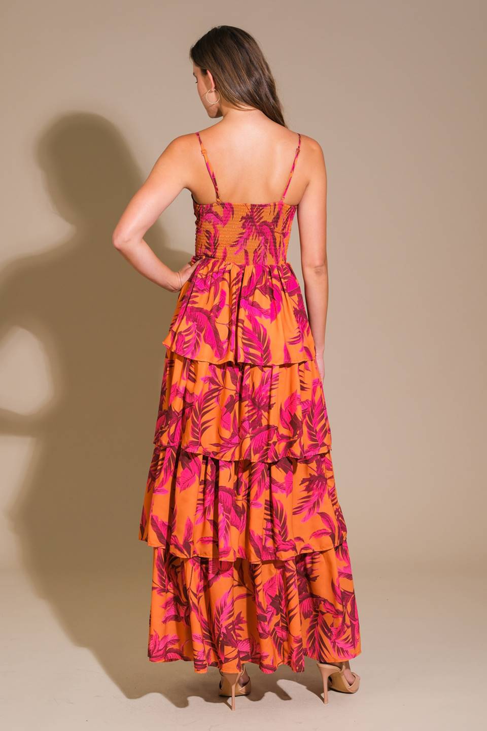 A woman is standing with her back to the viewer, wearing a colorful, tiered dress with orange and pink floral patterns. The background is a smooth, neutral beige.