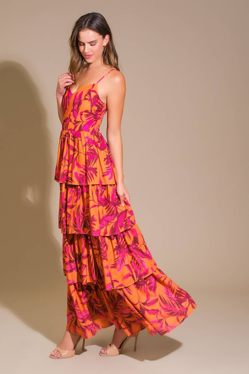 A woman is standing gracefully in a vibrant, tiered sundress featuring a floral pattern in orange and pink. The backdrop is a soft, neutral color, emphasizing the dress's lively colors.