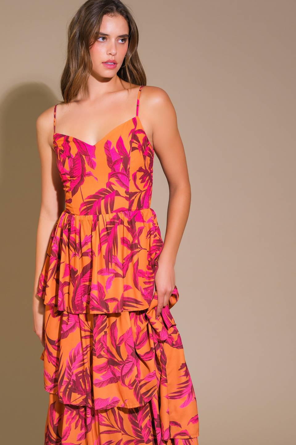 A model wears a vibrant, layered orange dress with pink tropical leaf patterns, standing against a neutral background, exuding a stylish and playful summer vibe.