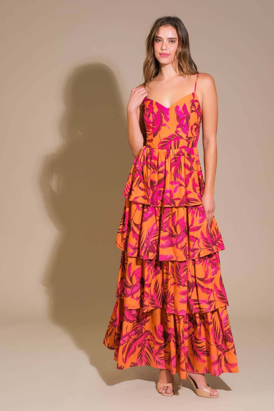 A model stands confidently wearing a vibrant, tiered dress with a mix of pink and orange colors, featuring a leafy print, against a neutral beige background.