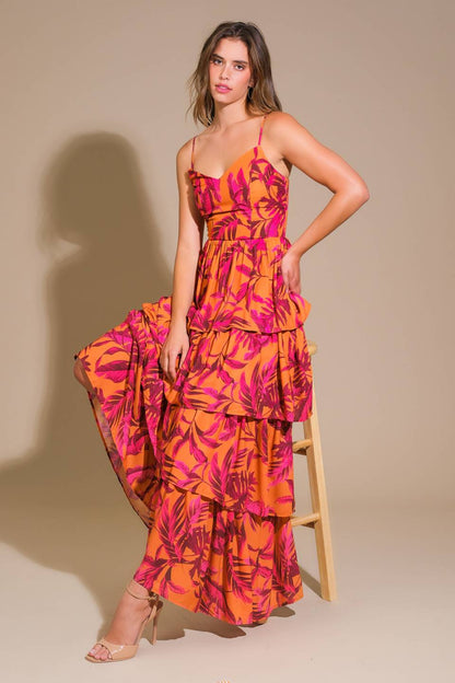 A woman sits on a wooden stool, wearing a vibrant, layered dress with pink and orange floral patterns. She poses in front of a neutral, beige background, exuding a confident demeanor.