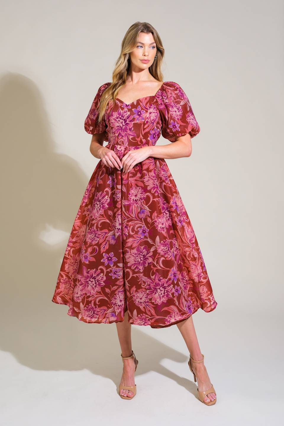 A woman stands confidently in a floral, burgundy dress with puffed sleeves, displaying graceful posture and styled hair against a neutral backdrop that emphasizes the vibrant patterns of her attire.