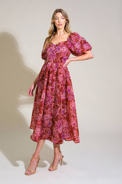 A woman stands confidently in a floral brown and pink dress with puffed sleeves, posing against a light beige background, showcasing a stylish outfit complemented by beige high-heeled sandals.