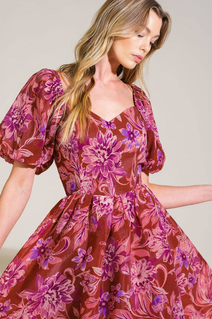 A woman wears a floral dress with puffed sleeves, gracefully looking down as she gently twirls. The context features a neutral background, emphasizing the vibrant patterns of her attire.