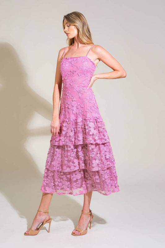 A model stands gracefully in a tiered, pink floral lace dress, with one hand on her hip. Soft lighting illuminates a minimalistic background, enhancing the dress's texture and color.