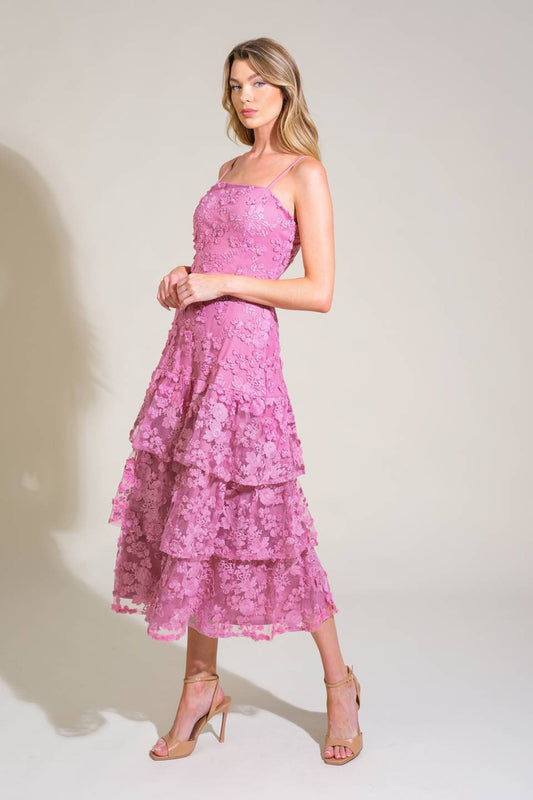 A woman stands gracefully in a layered pink lace dress with spaghetti straps, holding her arms at her waist, against a soft neutral background, showcasing elegance and style.