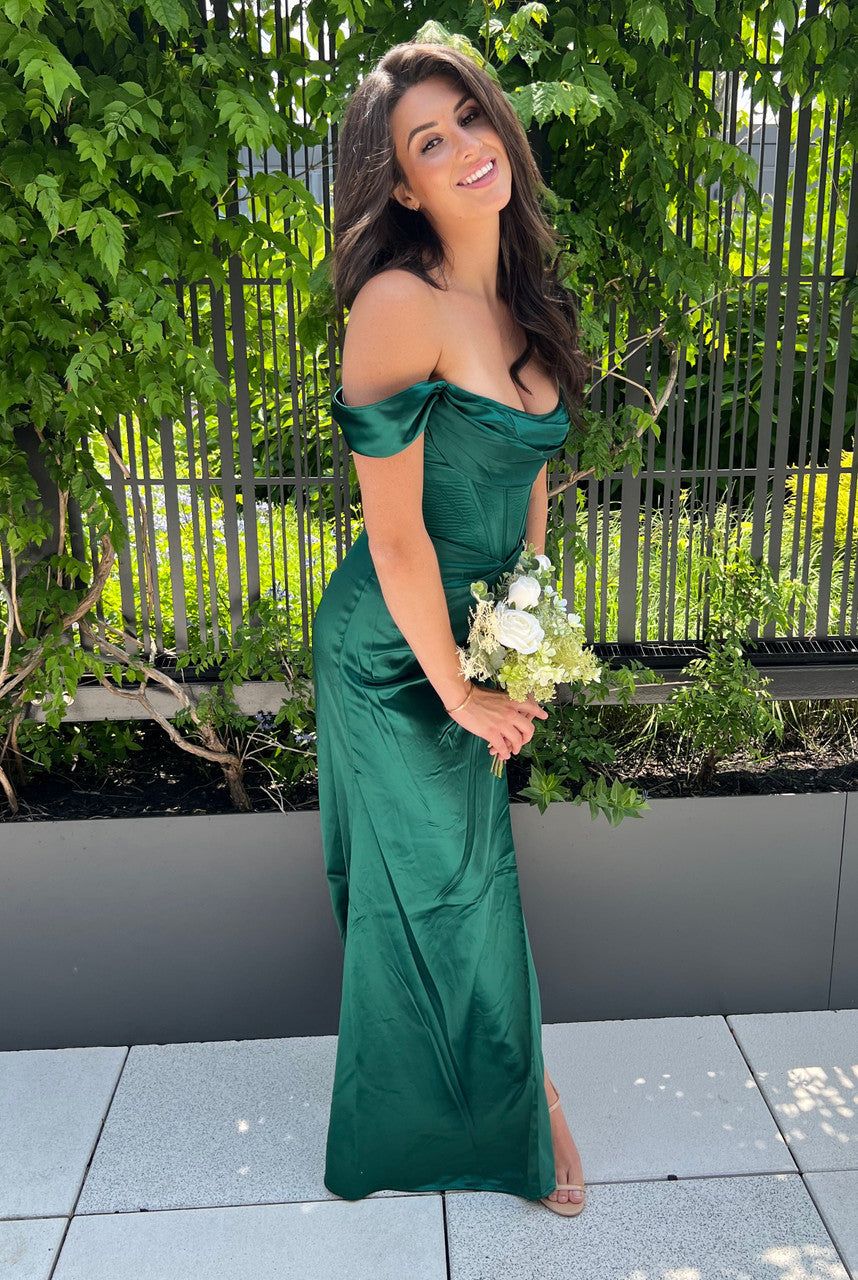 A woman in a green off-shoulder gown holds a bouquet of white and green flowers, smiling while standing in a lush outdoor setting with greenery and modern landscaping.